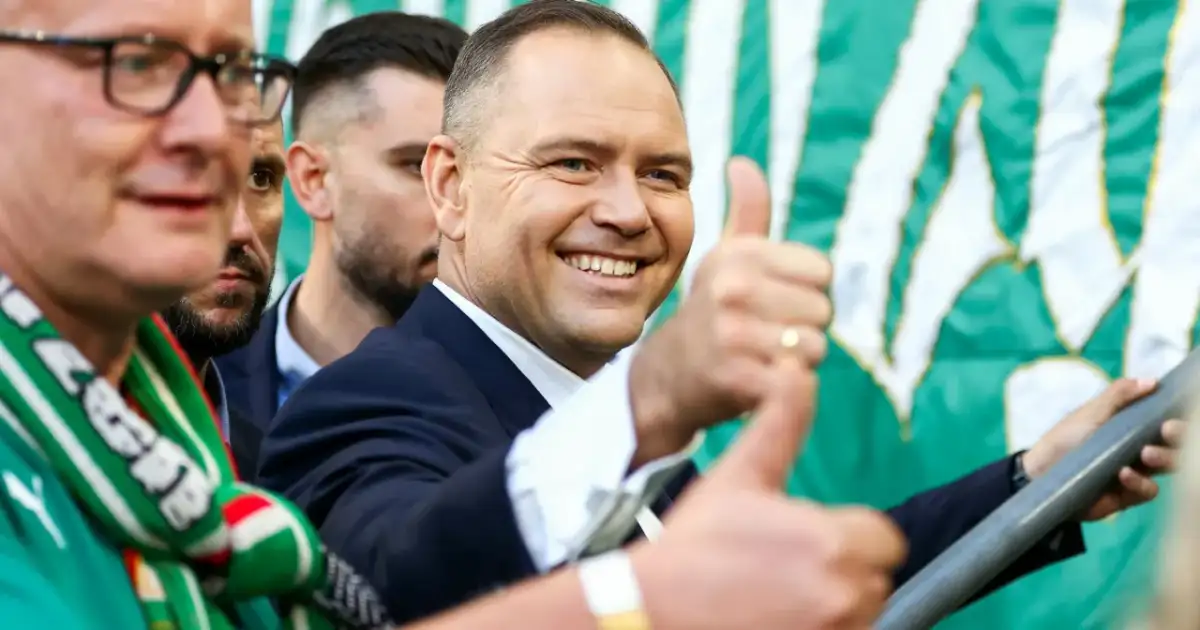 Uśmiechnięty mężczyzna w garniturze, z kciukiem w górze, na tle zielono-białej flagi. Widać kibiców Lechii Gdańsk.