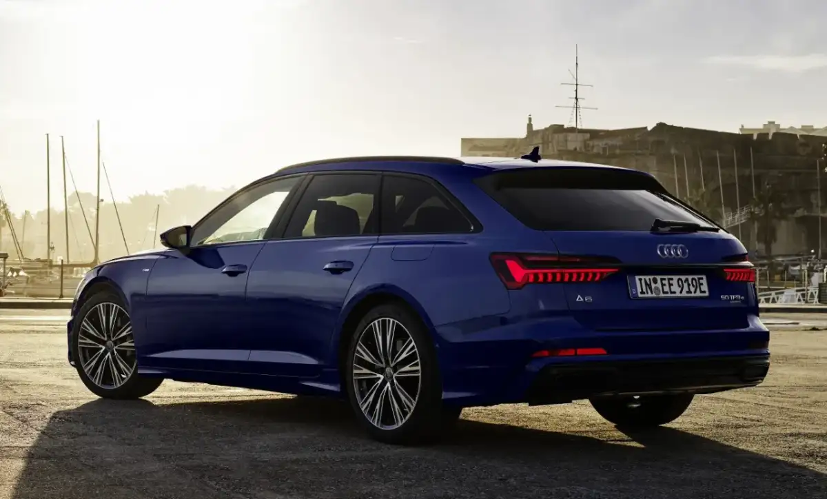 Niebieskie Audi A6 Avant z napisem "50 TFSI e" na klapie bagażnika, zaparkowane przy nabrzeżu z widokiem na zabytkową fortecę i żaglówki.