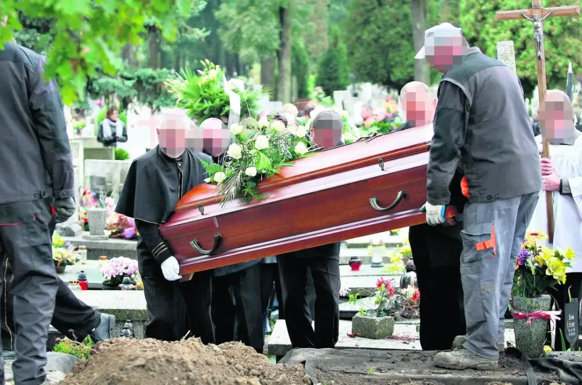 Pogrzeb na cmentarzu. Kilku mężczyzn niesie drewnianą trumnę ozdobioną kwiatami w kierunku grobu głębinowego.