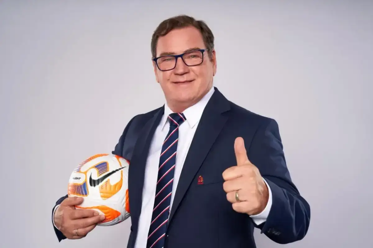 Uśmiechnięty trener reprezentacji Polski w garniturze trzyma piłkę i pokazuje kciuk w górę.