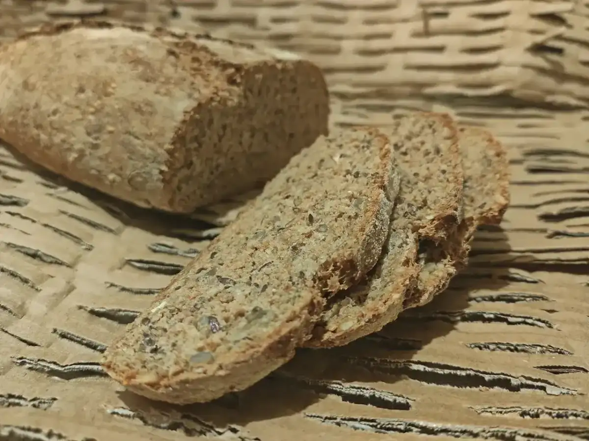 Chleb orkiszowy, pokrojony na kromki, z widocznymi ziarnami, leży na falistej tekturze.
