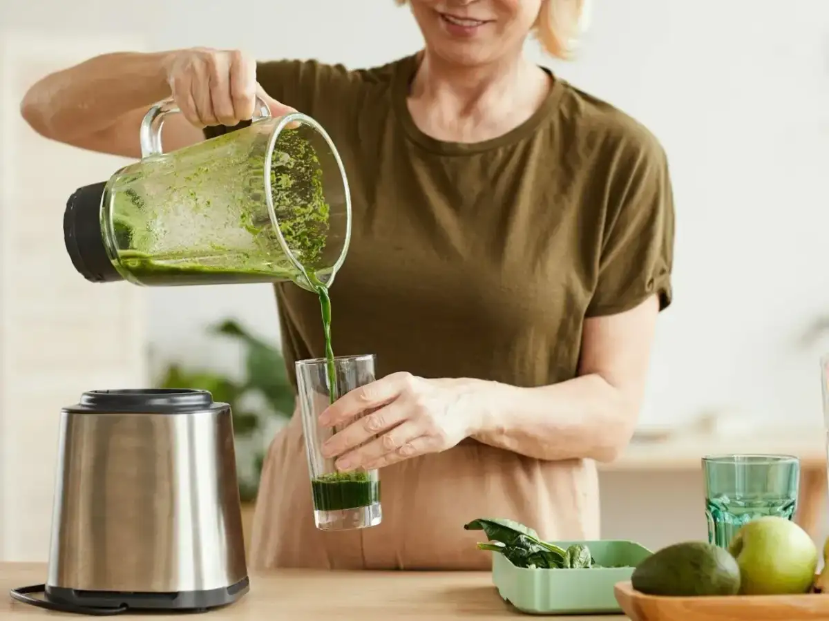 Kobieta nalewa zielony koktajl położnej do szklanki. Na blacie widać blender, awokado i jabłko.
