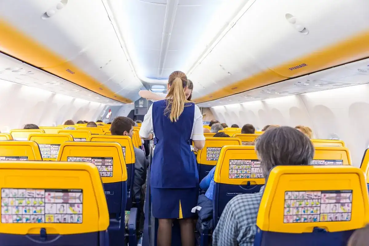 Wnętrze samolotu Ryanair, stewardessa obsługuje pasażerów. Żółte zagłówki siedzeń i pomarańczowe pasy na suficie tworzą słoneczną atmosferę.