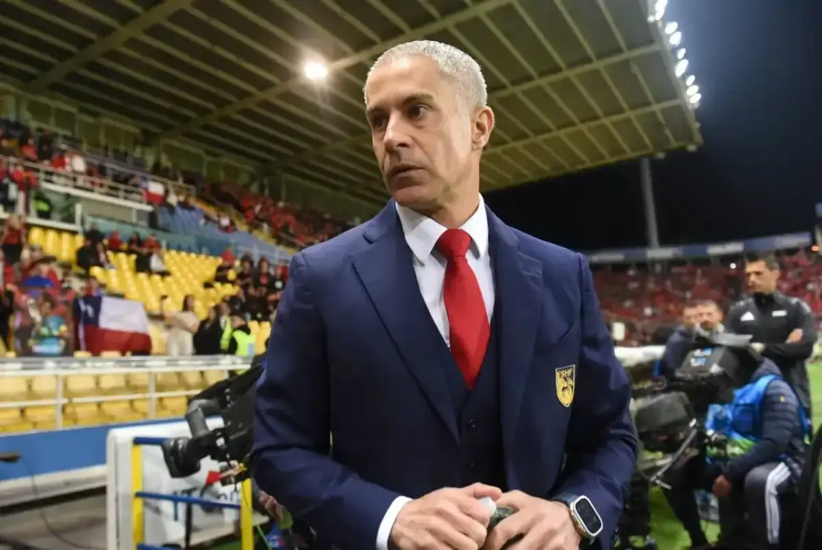 Sylvinho, trener w granatowym garniturze i czerwonym krawacie, stoi na tle trybun stadionu z flagą Chile.