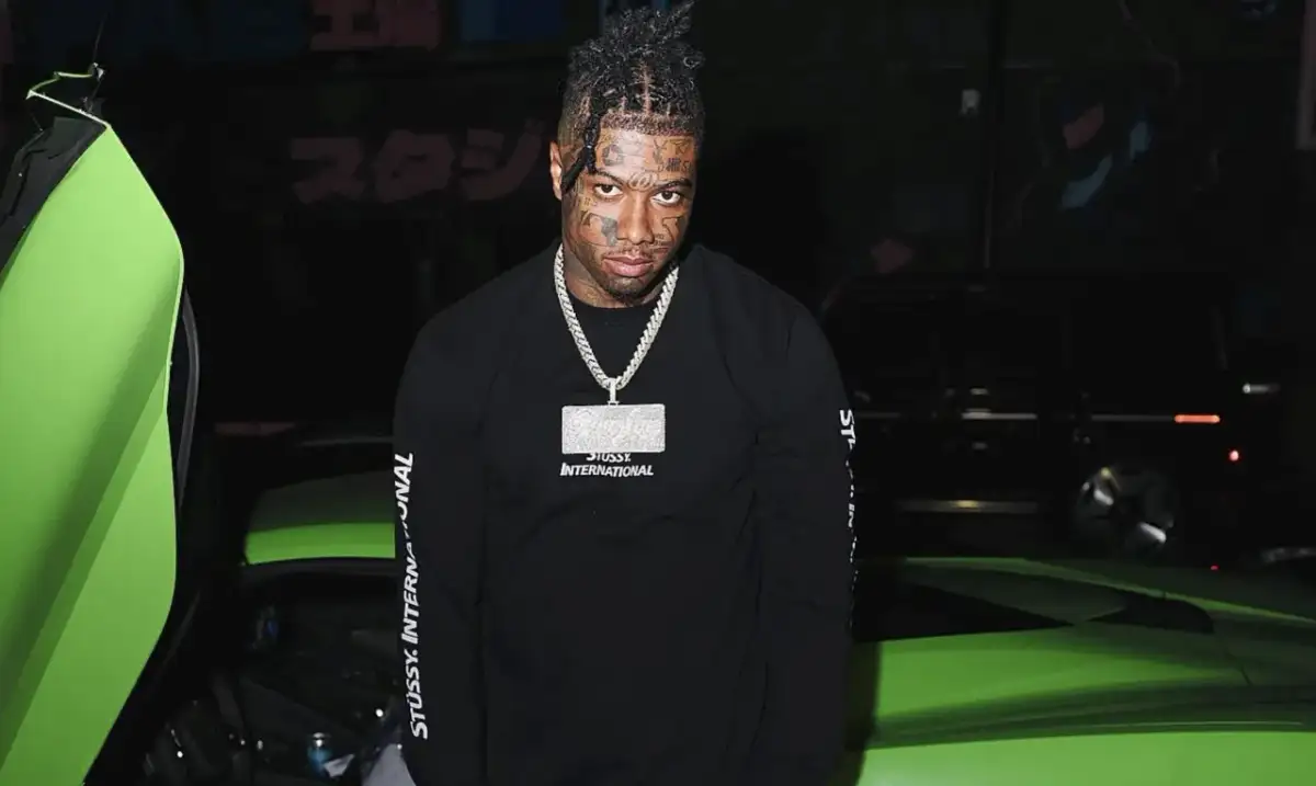 Raper z tatuażami na twarzy, znany jako blueface, pozuje obok jaskrawozielonego samochodu sportowego.