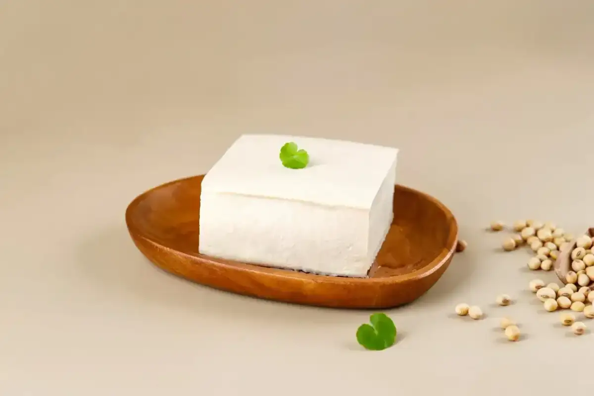Kostka białego tofu na drewnianej miseczce, ozdobiona listkiem. Obok garść ziaren soi. To właśnie co to tofu – produkt z soi.