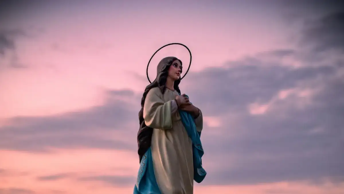 Figurka Matki Boskiej z aureolą na tle różowego nieba. W dłoniach trzyma błękitną szatę, jakby w modlitwie "Anioł Pański".