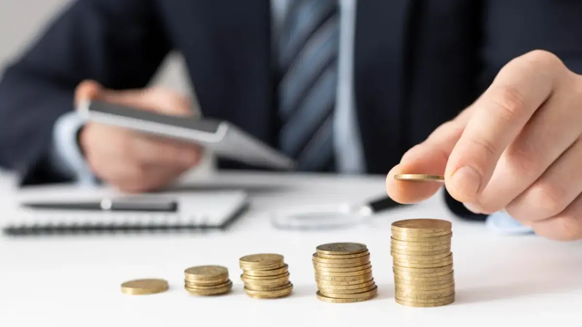 Dłoń układa monety, budując stosy. W tle mężczyzna z kalkulatorem, sugerującym planowanie finansowe i poszukiwanie optymalnych rozwiązań, być może związanych z **rajami podatkowymi**.