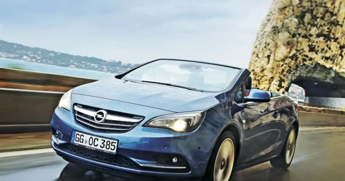 Niebieski Opel Cascada z opuszczonym dachem jedzie po mokrej drodze przy skalistym wybrzeżu.