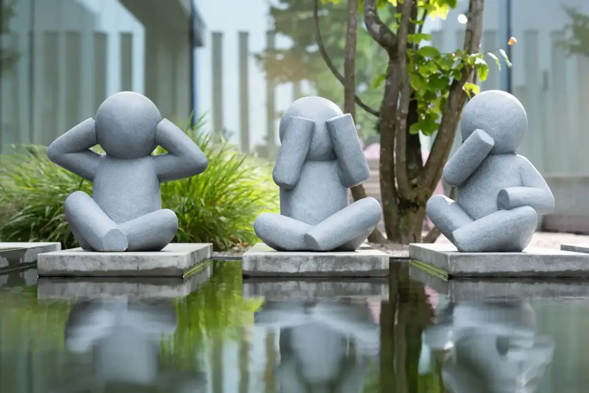 Drei graue Skulpturen von sitzenden Figuren, die ihre Ohren, Augen und Mund bedecken, spiegeln sich in einem Teich.