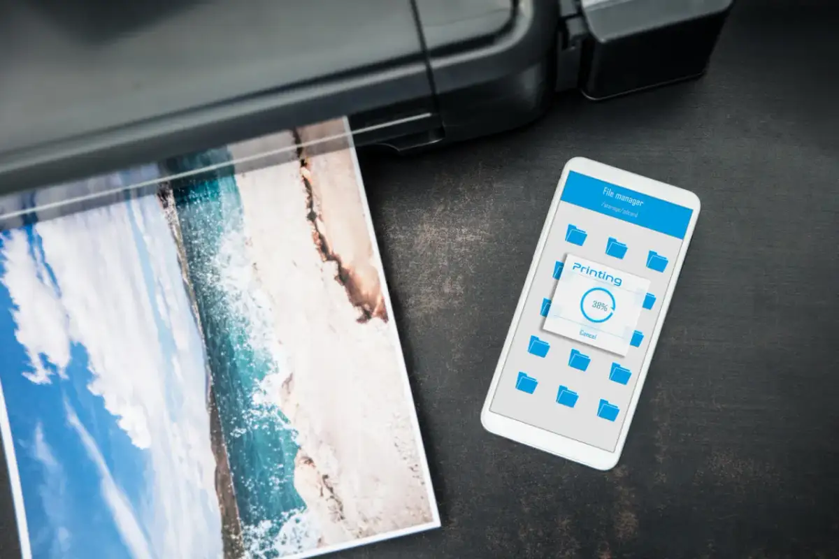 Jak wydrukowac cos z iPhone? Widok telefonu z ekranem "Printing 38%" obok drukarki drukującej zdjęcie plaży.