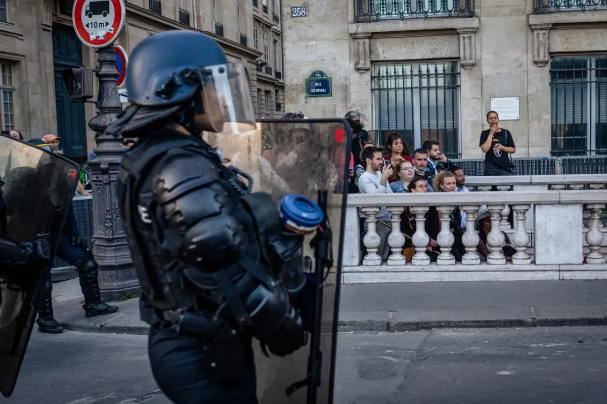 Policjant w pełnym rynsztunku ochronnym z tarczą, obserwuje tłum za barierką.