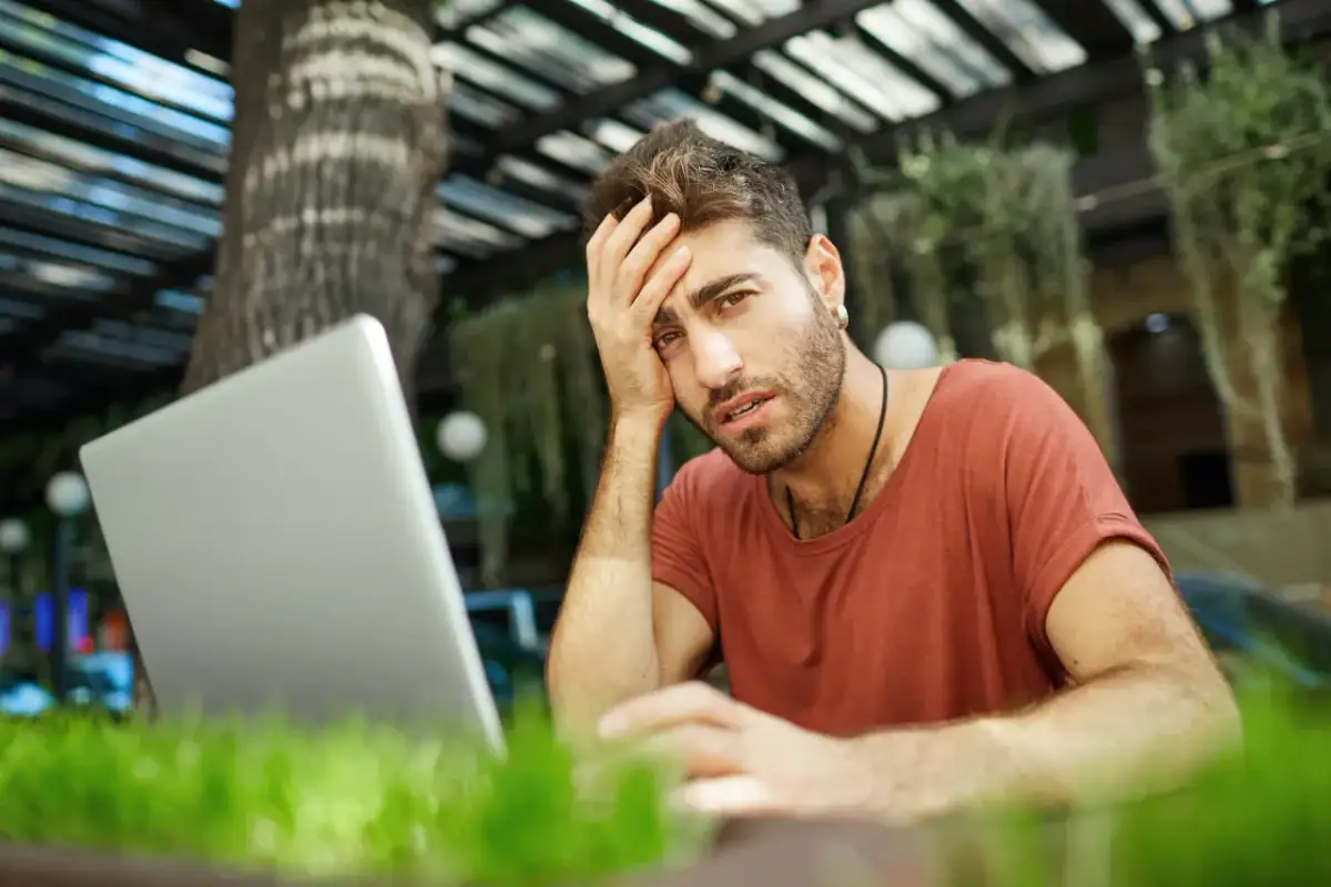 Mężczyzna z ręką na czole, sfrustrowany pracą przy laptopie. Czyżby padł ofiarą pomówienia w internecie?