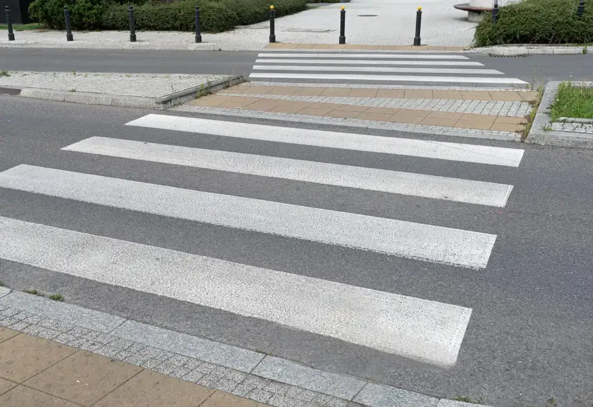 Sugerowane przejście dla pieszych z białymi pasami na szarym asfalcie, prowadzące przez chodnik i trawnik.