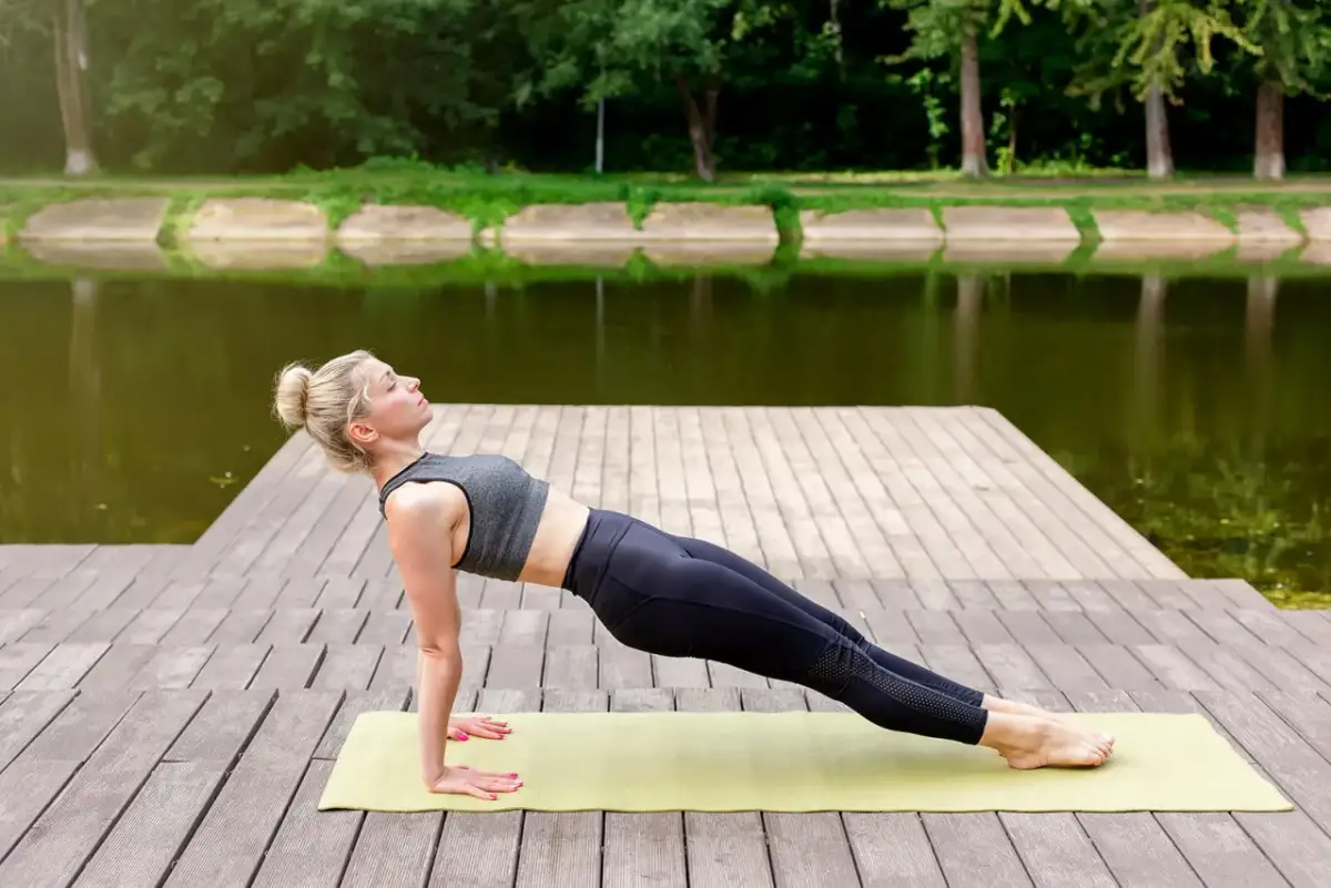 Kobieta wykonuje ćwiczenie reverse plank na macie na drewnianym pomoście nad jeziorem.