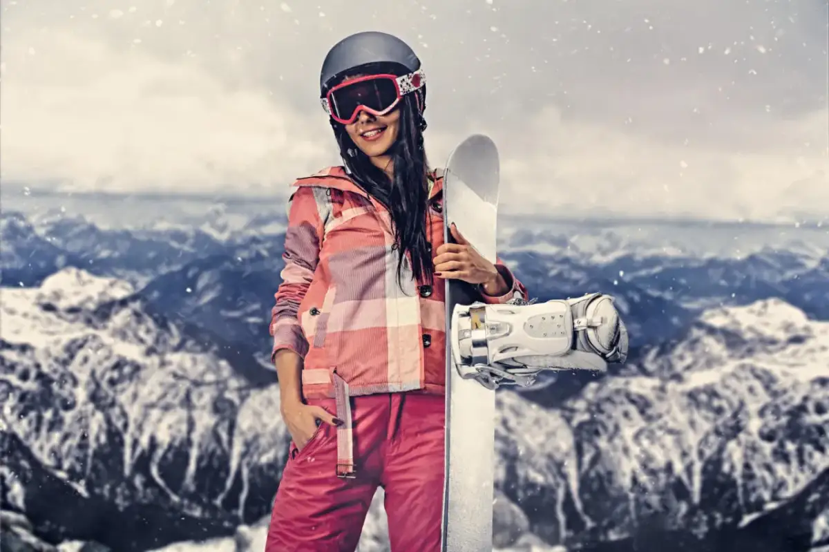 Kobieta w stroju narciarskim trzymająca snowboard na tle gór.