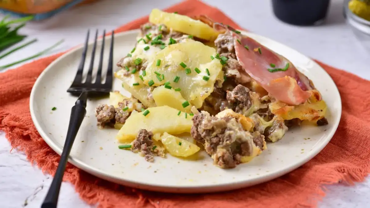 Zapiekanka ziemniaczana z mięsem mielonym, posypana szczypiorkiem, na talerzu z widelcem.
