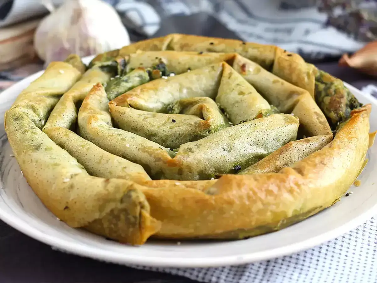 En gyllene, spiralformad burek med spenatfyllning, perfekt för ett hemlagat burek recept. Vitlök och lök i bakgrunden.