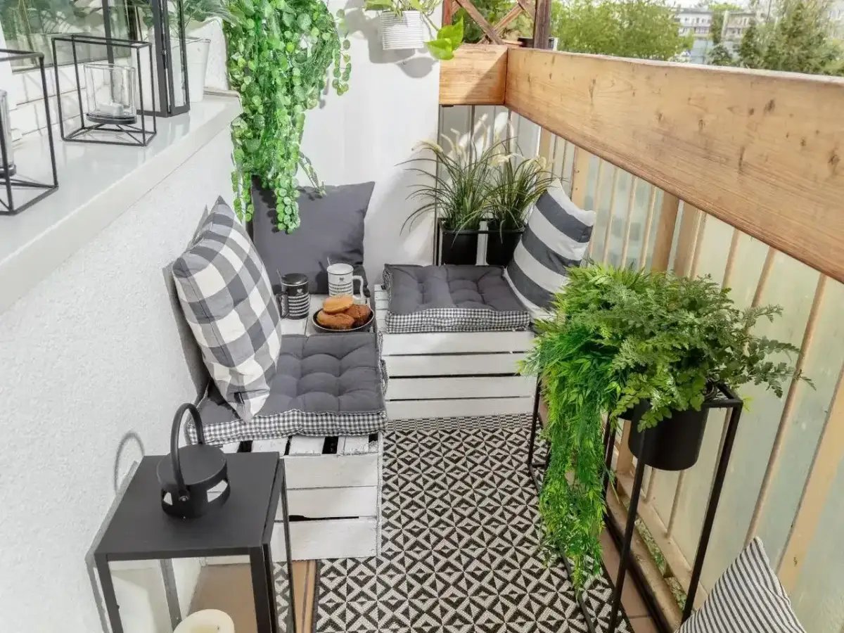 Przytulny balkon w bloku z wygodnymi siedziskami z palet, mnóstwem zieleni i czarno-białymi dodatkami. Idealne miejsce na poranną kawę.