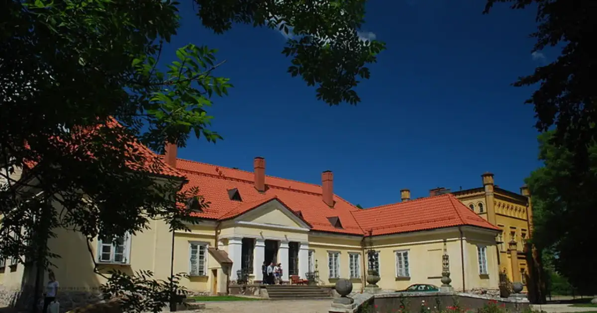 Pałac w Waplewie z czerwonym dachem i żółtymi ścianami, otoczony zielenią, pod błękitnym niebem.