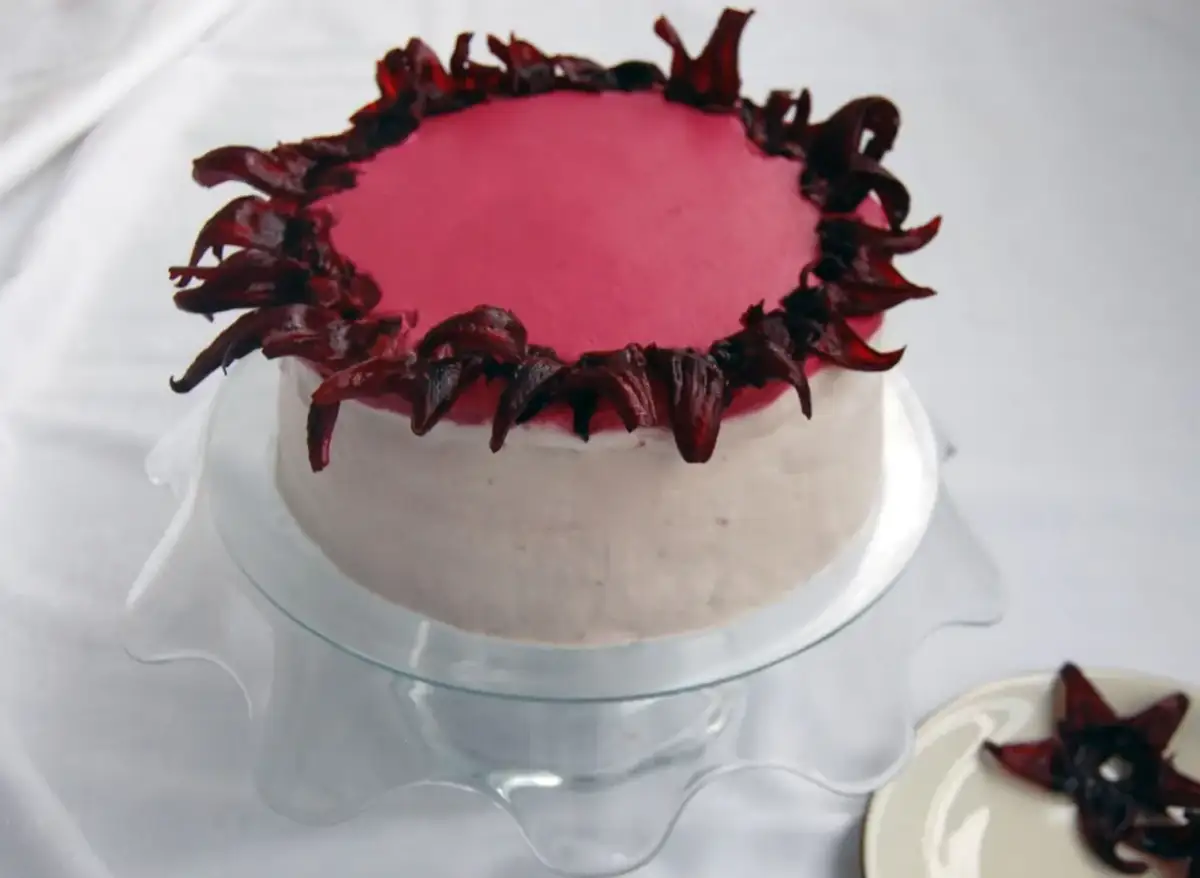 Różowy tort udekorowany ciemnoczerwonymi płatkami hibiskusa na górze i bokach.
