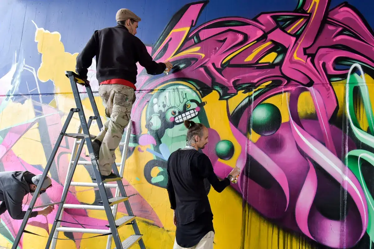 Drei Künstler sprühen bunte Graffiti auf eine Wand, einer auf einer Leiter.