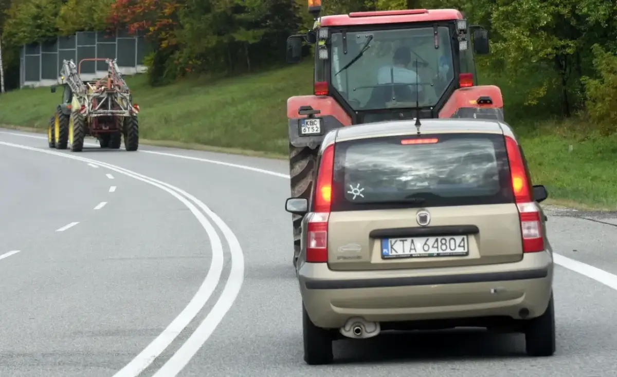 Złoty Fiat Panda jedzie za czerwonym traktorem. Czy można wyprzedzić traktor na podwójnej ciągłej? Droga kręta, drzewa po bokach.