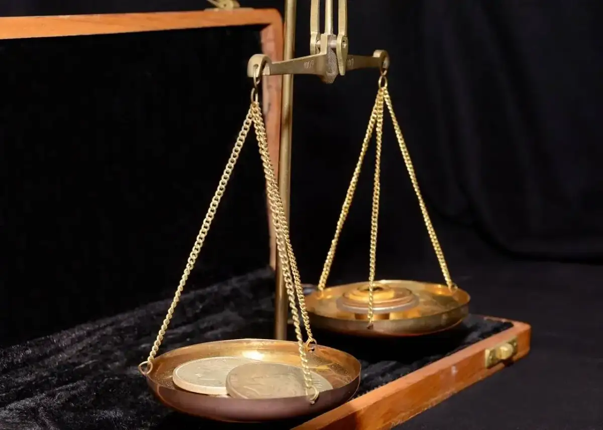 Waga szalkowa z monetami i ciężarkami. Na jednej szalce leżą monety, na drugiej ciężarki, symbolizujące uncję złota.