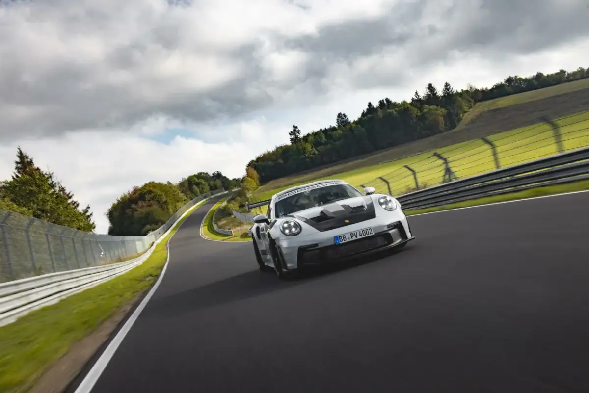 Biały Porsche 911 GT3 RS z czarnymi akcentami pędzi po torze wyścigowym wśród zielonych wzgórz i chmur.