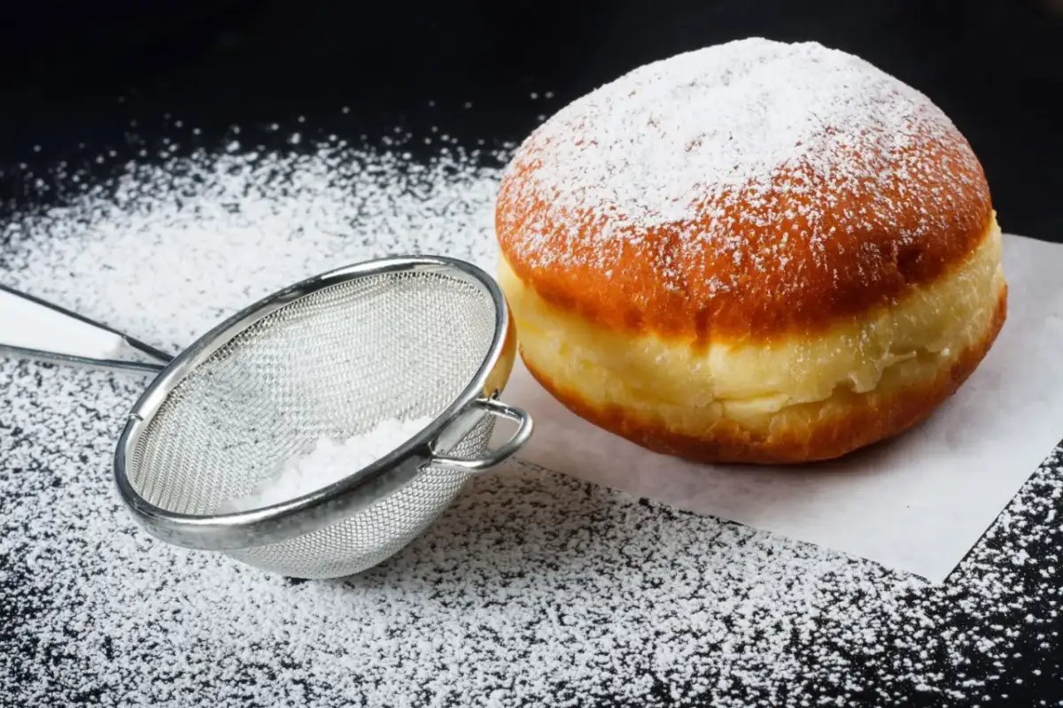 Puszysty pączek posypany cukrem pudrem, obok sitko z cukrem. Idealne na tłusty czwartek, pyszne pączki!