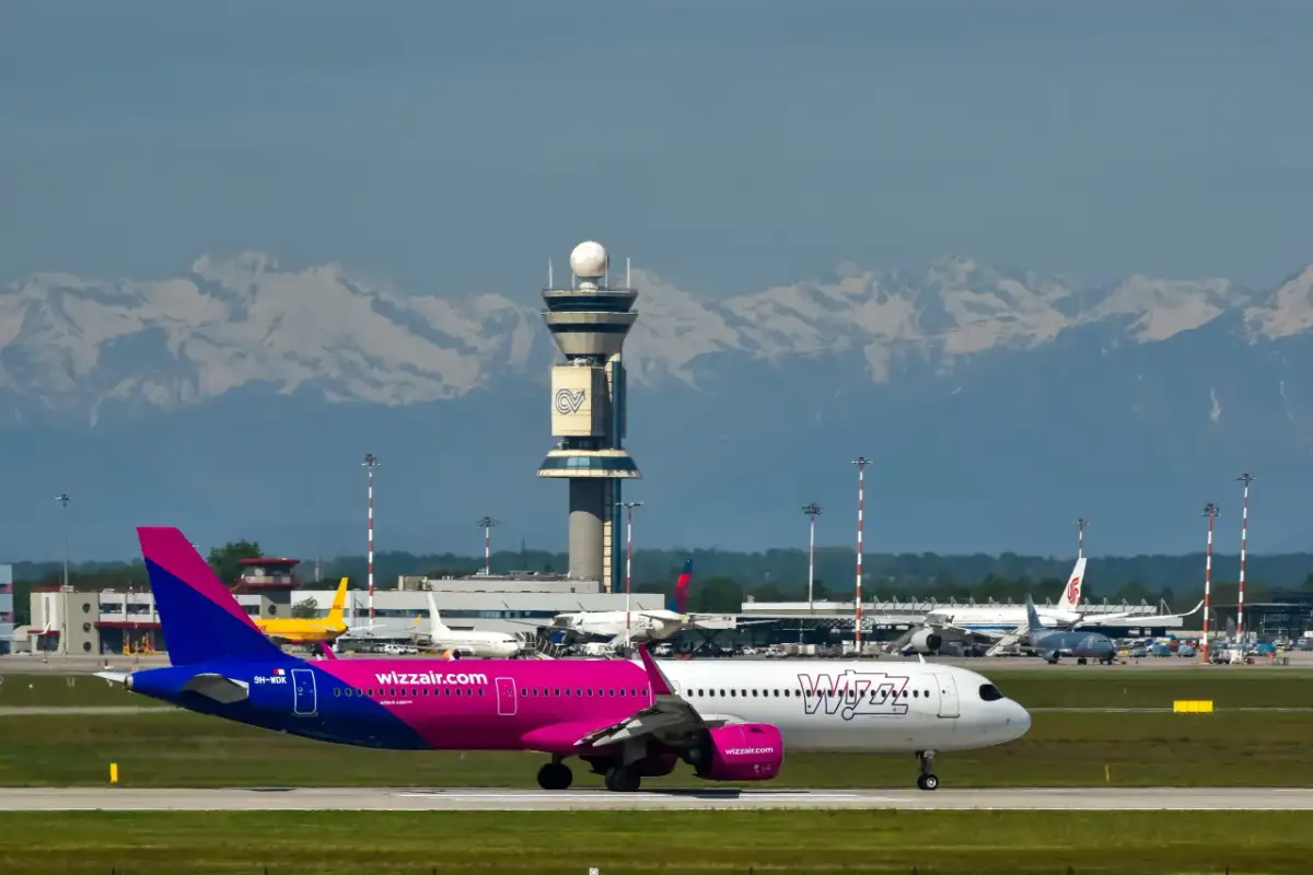 Samolot Wizz Air na płycie lotniska z wieżą kontroli lotów i górami w tle.
