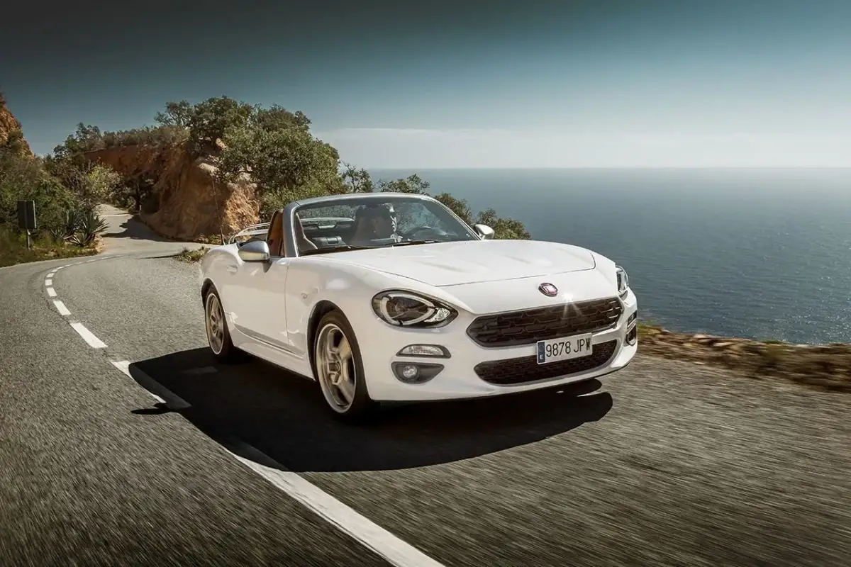 Biały Fiat 124 Spider sunie po krętej drodze nad morzem.