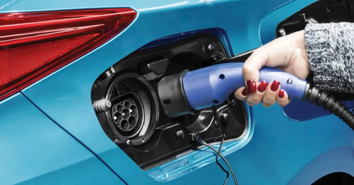 Dłoń z czerwonym lakierem na paznokciach podłącza ładowarkę do niebieskiej hybrydy plug-in.