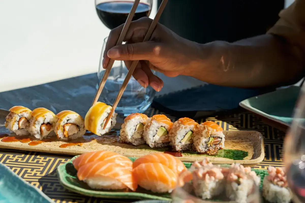 Ręka z pałeczkami sięga po sushi. Na talerzach różne rodzaje roladek i nigiri. Ile sztuk sushi na osobę? Trudno ocenić, ale wygląda apetycznie.