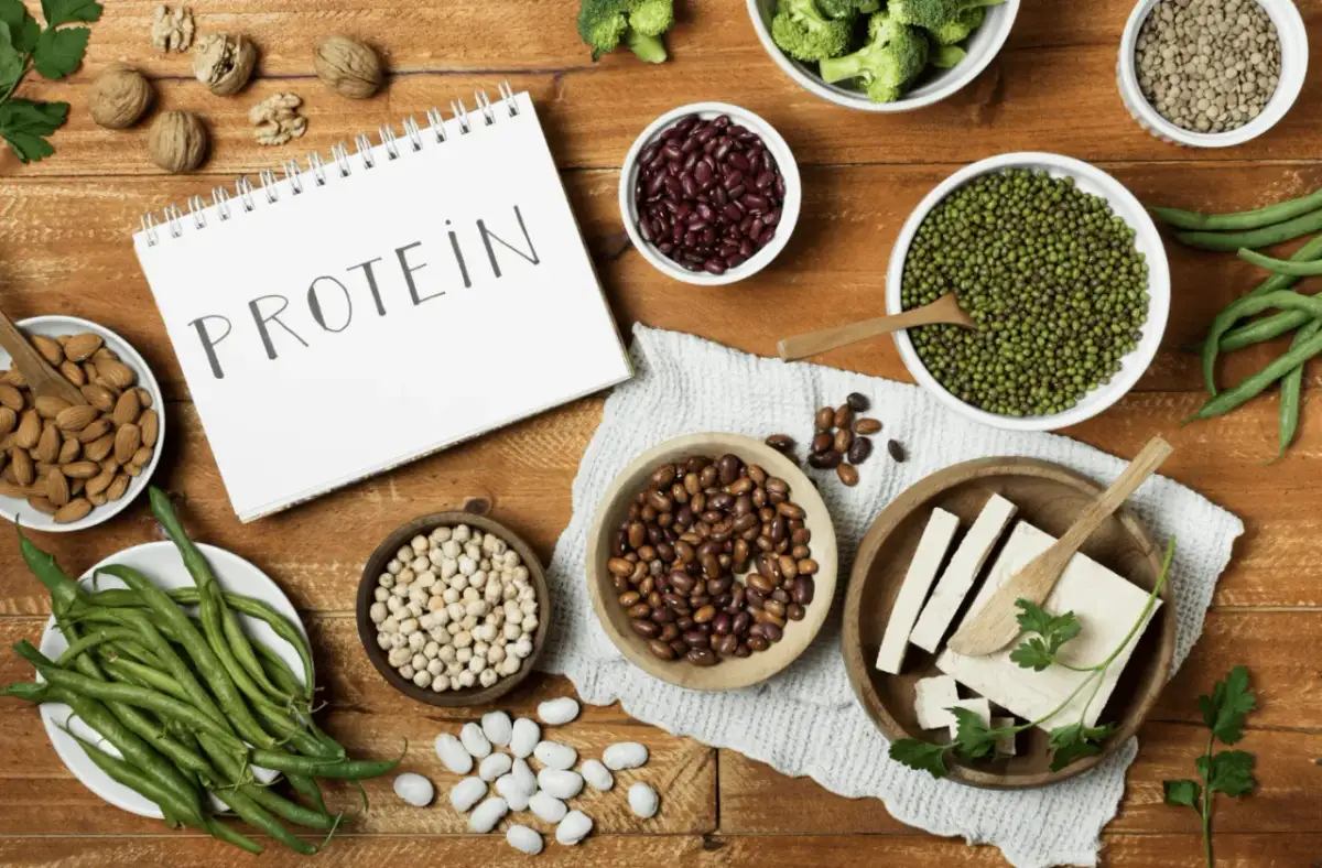 Notatnik z napisem "PROTEIN" otoczony miseczkami z fasolą, ciecierzycą, soczewicą, orzechami, brokułami, fasolką szparagową i tofu.