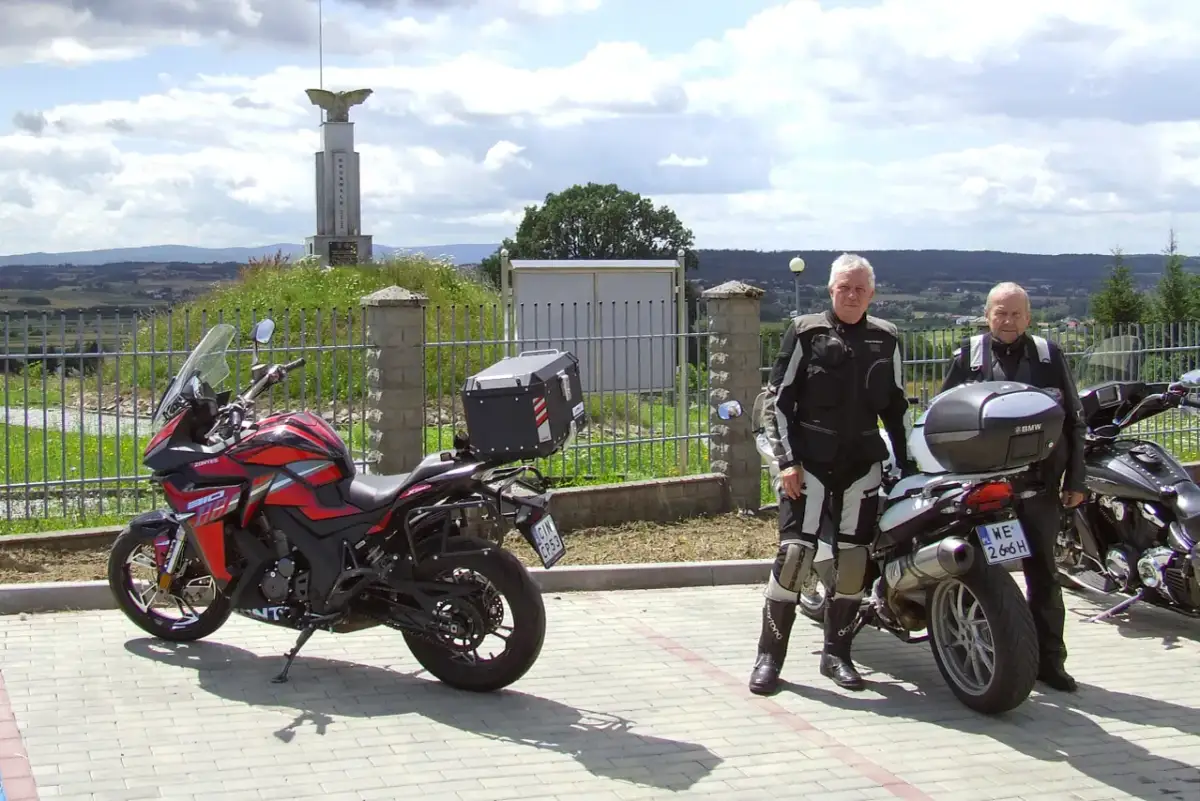 Dwóch motocyklistów w strojach turystycznych stoi obok swoich motocykli na tle krajobrazu i pomnika.
