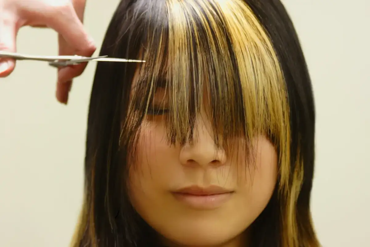 Fryzjerka przycina grzywkę w stylu birkin bangs. Dwukolorowe pasma opadają na twarz.