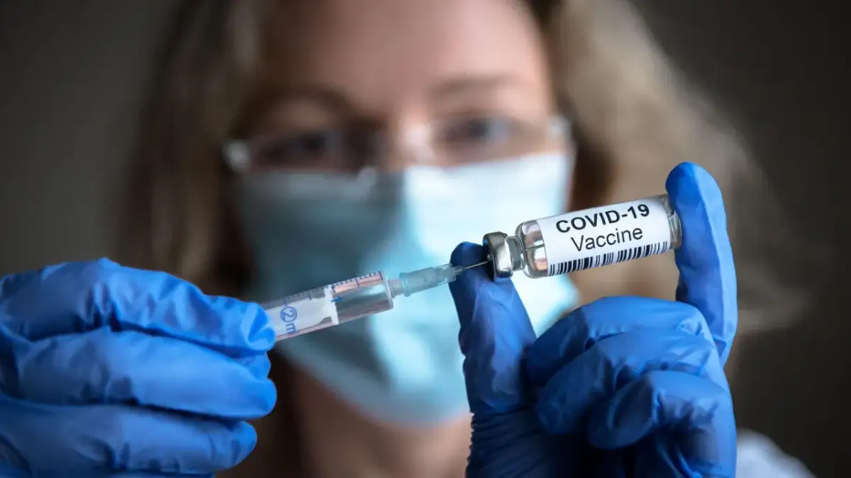 Osoba w niebieskich rękawiczkach przygotowuje szczepionki na covid, pobierając płyn do strzykawki z fiolki z napisem "COVID-19 Vaccine".