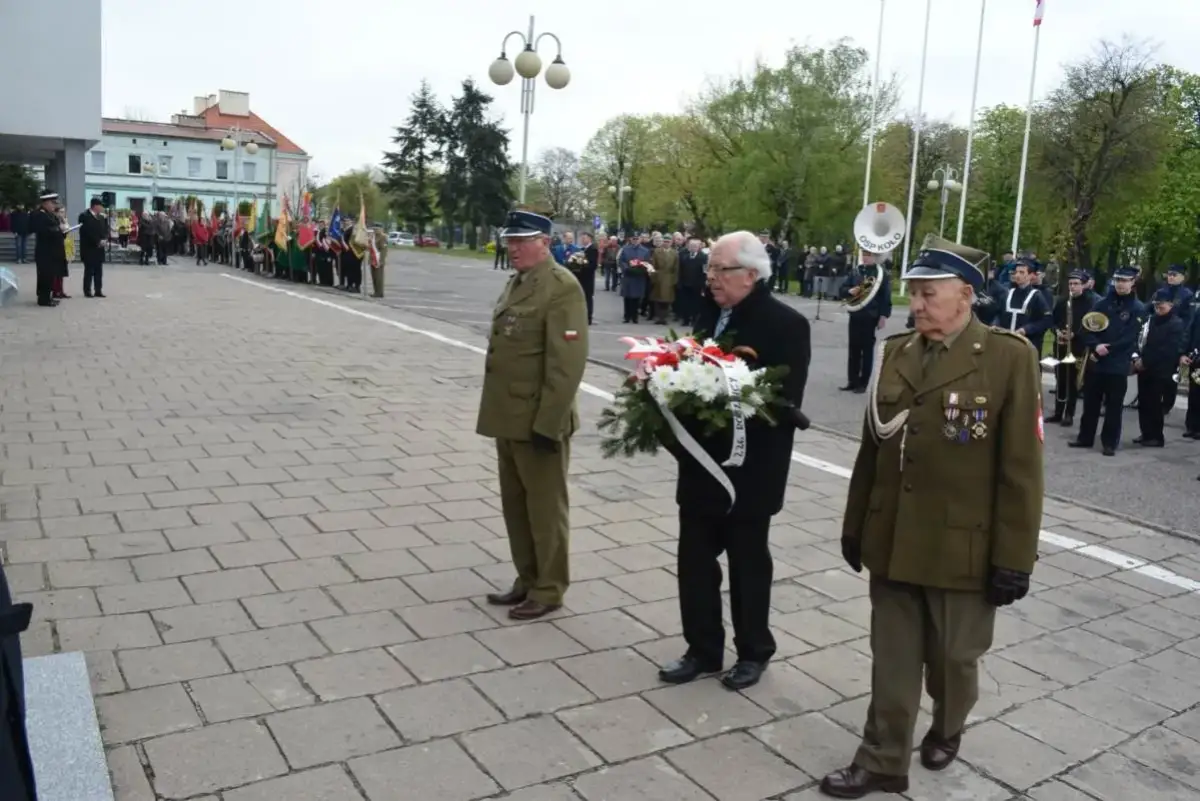 Trzech mężczyzn w mundurach wojskowych i cywilnych, jeden z nich niesie wieniec. W tle parada i orkiestra.