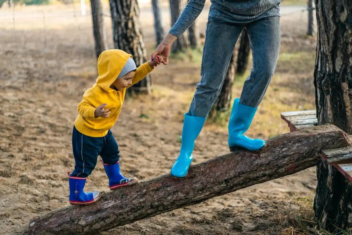 Dziecko w żółtej bluzie i mama w niebieskich kaloszach idą po kłodzie. Czy po szczepieniu można iść z dzieckiem na spacer? Tak, jeśli czuje się dobrze!