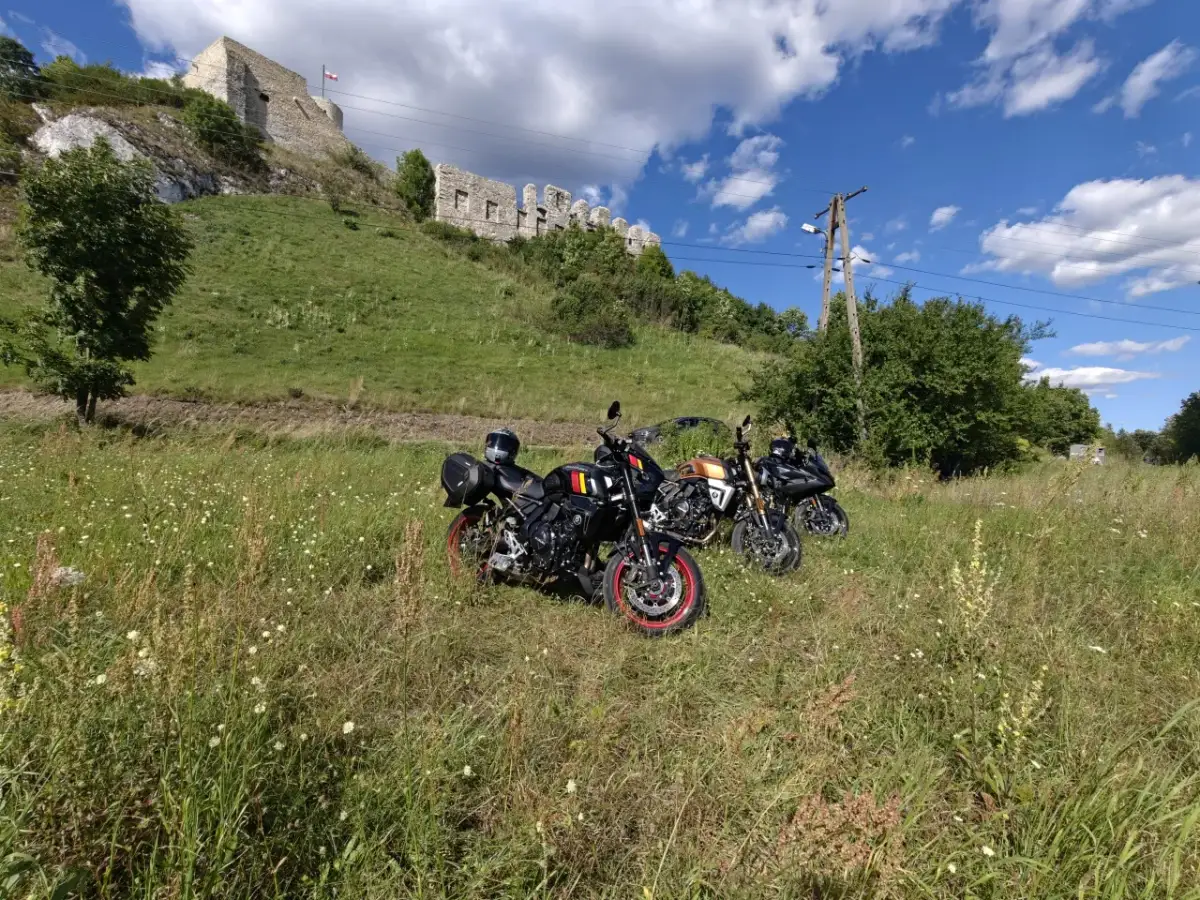 Trzy motocykle stojące na łące z widokiem na ruiny zamku na wzgórzu.