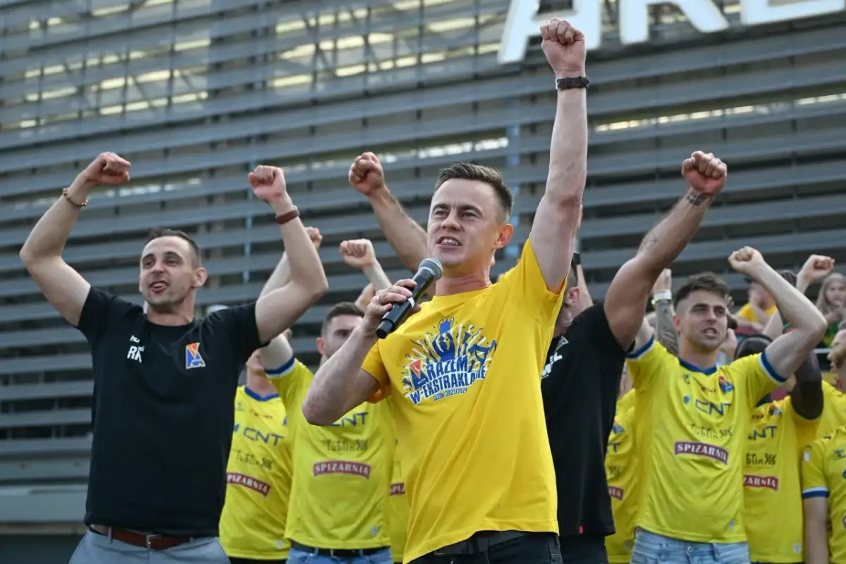 Mężczyzna w żółtej koszulce z mikrofonem i kibice z podniesionymi pięściami.