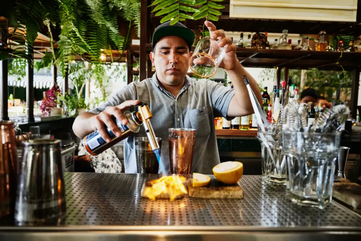 Barman podgrzewa szkło palnikiem na tle baru z butelkami i roślinami.