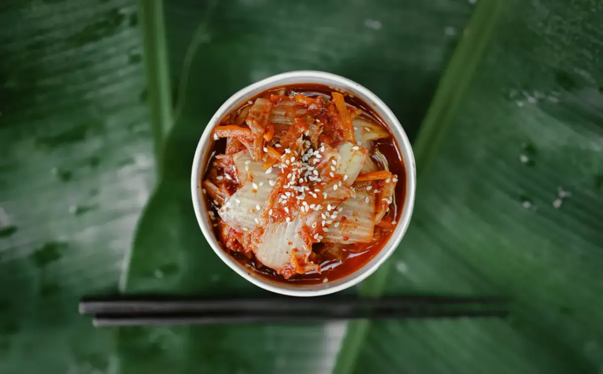 Kapusta kimchi co to jest? Pikantna, fermentowana kapusta pekińska z sezamem, podana w białym naczyniu na tle zielonych liści. Obok leżą pałeczki.