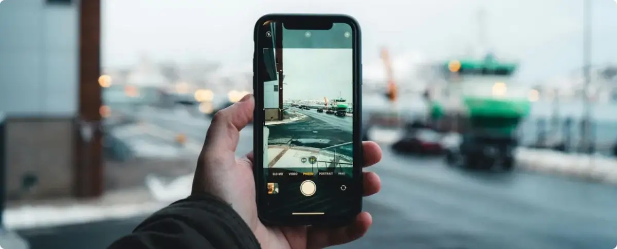 Dłoń trzyma smartfon, na ekranie którego widać interfejs aparatu. Pokazuje, jak dziala aparat, rejestrując zimowy krajobraz z drogą i zielonym statkiem.