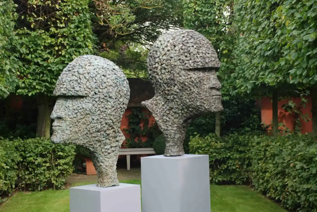 Zwei moderne Gartenskulpturen in Form von Köpfen aus Stein, im Grünen auf Podesten platziert.