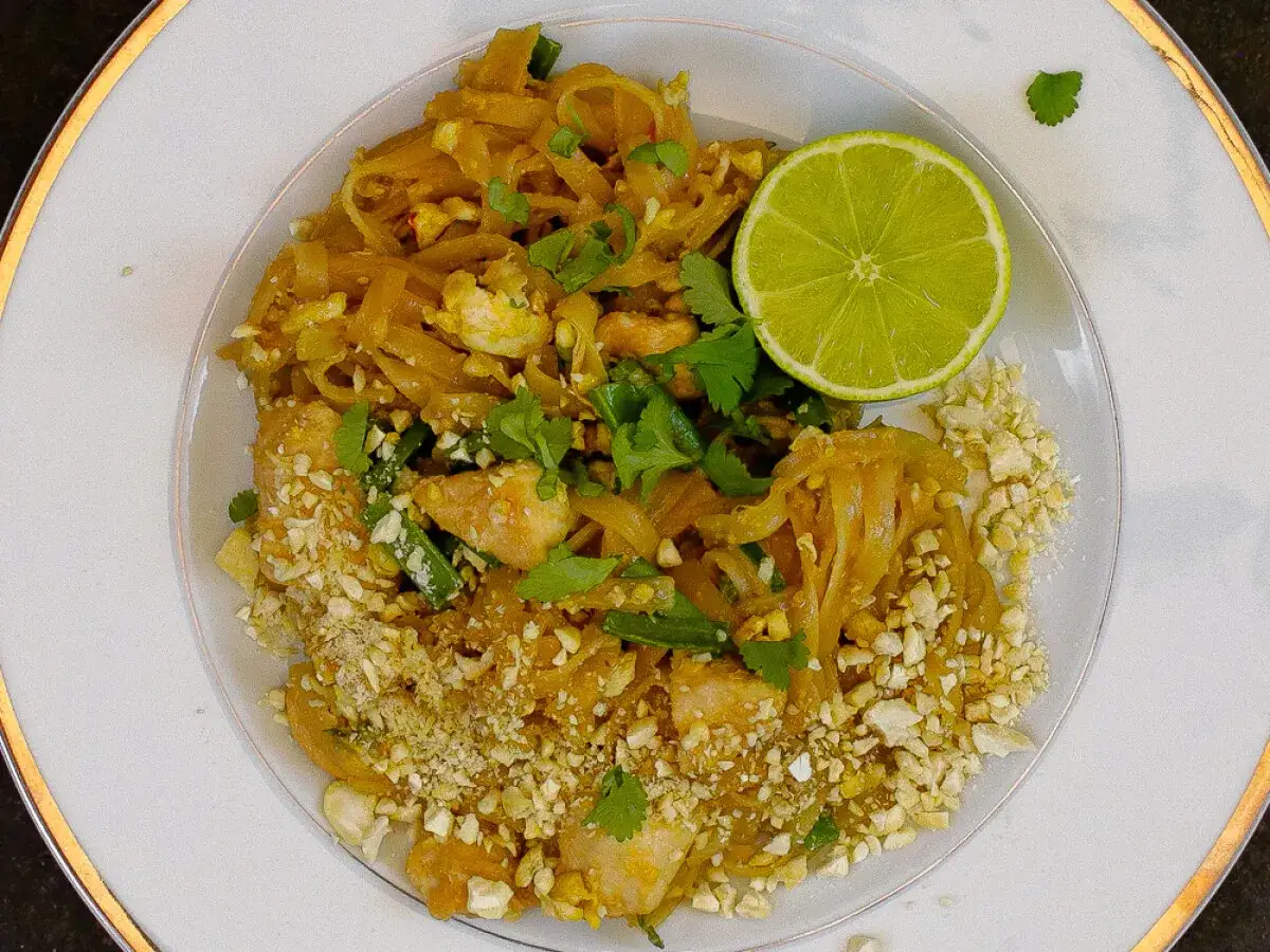 Talerz z Pad Thai, posypany orzeszkami ziemnymi, z połówką limonki i kolendrą.