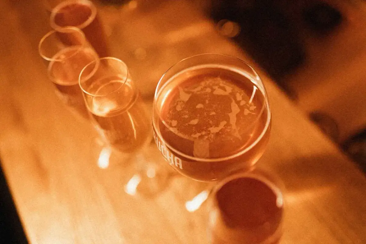 Kilka kieliszków z bursztynowym napojem, jeden z nich z widocznym napisem "kombucha".