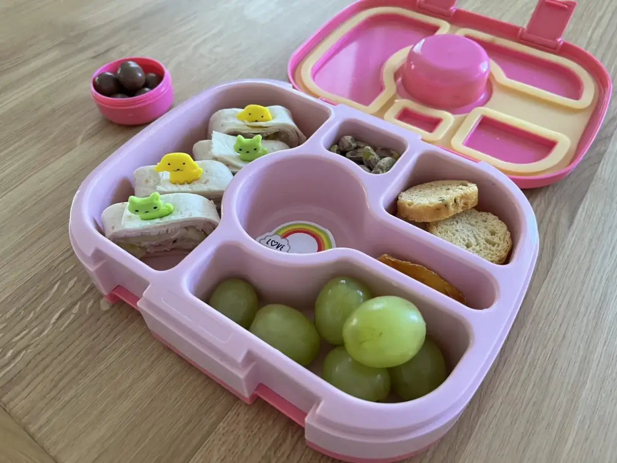Różowa śniadaniówka do szkoły z przegródkami zawiera zawinięte tortille z uroczymi ozdobami, winogrona, orzeszki pistacjowe, krakersy i suszone mango.