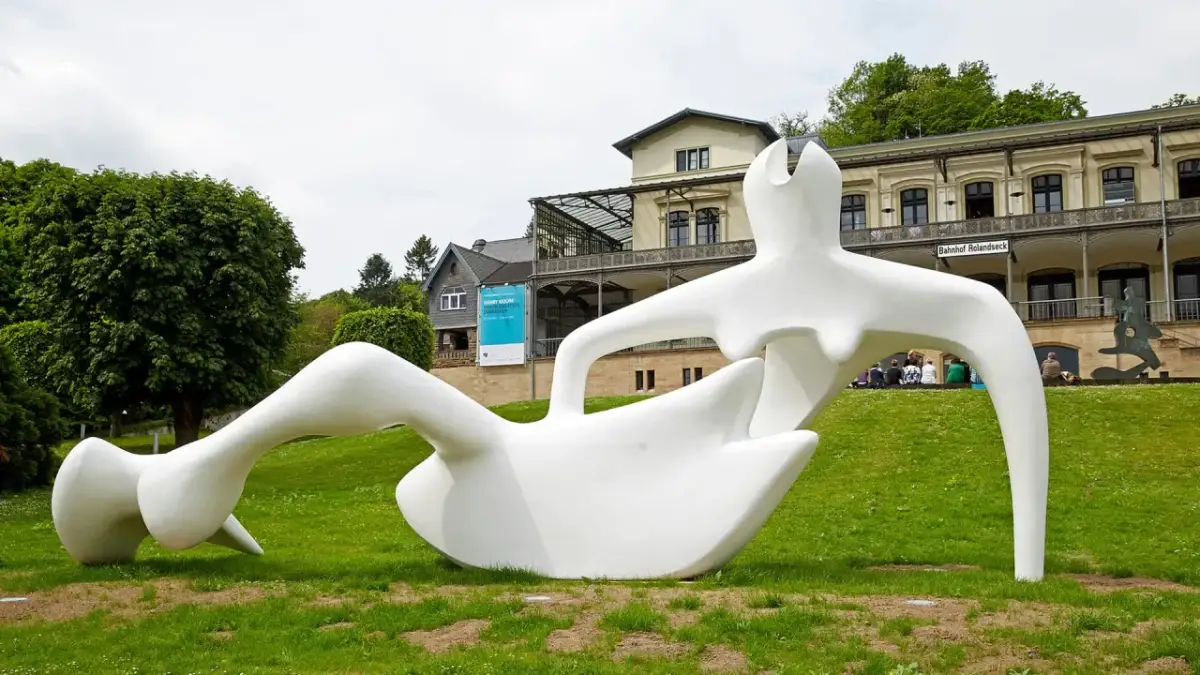 Weiße Skulptur einer liegenden Figur im Freien vor einem Gebäude mit der Aufschrift "Bahnhof Rolandseck".